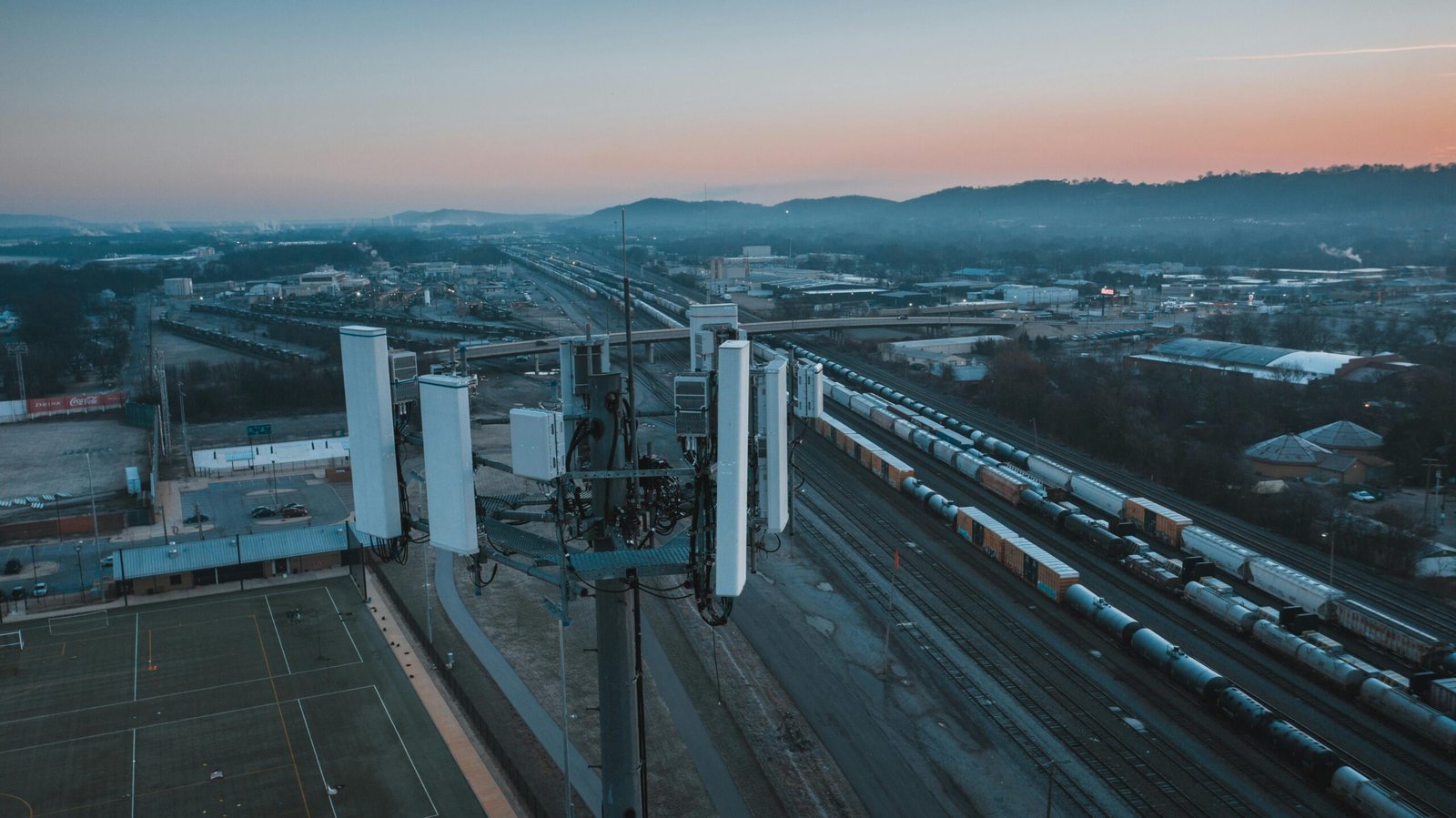 Aerial view of modern cell tower and railway tracks with cargo containers near rugby fields under sunset sky in town